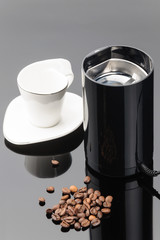 Coffee grinder with coffe beans and cup on the grey mirror background