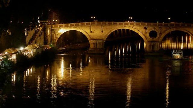 Timelapse Rome by night at Summer - Many people walk on the illuminated quays near the river with the Sisto bridge and the dome of St. Peter in the distance