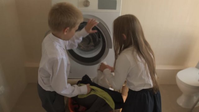 Back to school. Happy children with blue eyes and blond hair in school uniform. Boy and girl of preschool age load toys for washing in washing machine at home in bathroom. Shallow focus