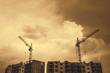 Cranes above unfinished multistorey panel building on background of cloudy sky with copy space in sepia tones. Process of construction of apartment building in overcast weather close up in monochrome.
