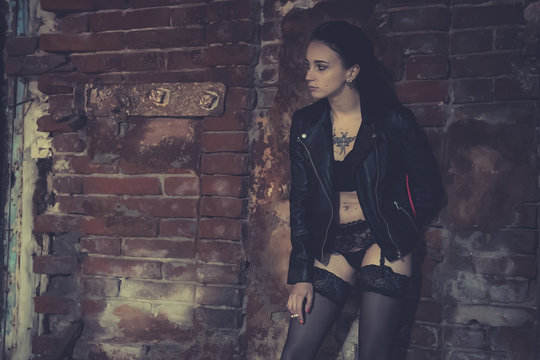Sexy model stands old brick wall and poses in the image of a corrupt girl. Prostitute on the street smoking a cigarette with his knee bent. Girl addict in a leather jacket.