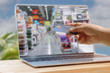 Hand holding mockup of credit card above laptop keyboard with image of supermarket on screen which is on wooden table outdoor with nature green background. Online shopping and ecommerce concept.