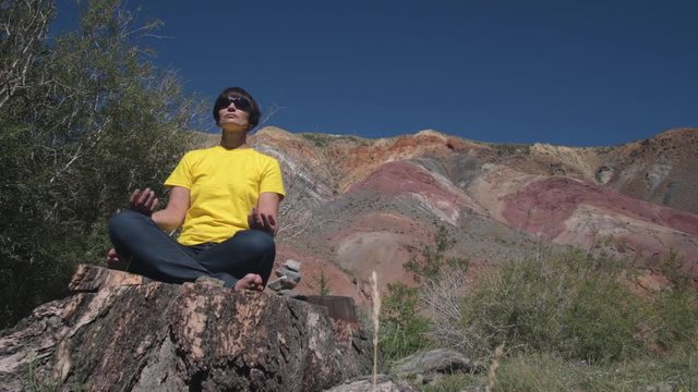 A female traveler sits on a huge tree stump and meditates against the beautiful mountains.