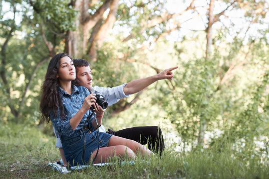 young couple sitting on the grass in the forest, taking photos and looking on sunset, summer nature, bright sunlight, shadows and green leaves, romantic feelings