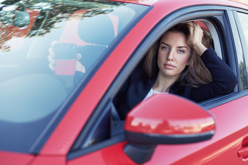 Gorgeous Caucasian fashionable female model in formal clothes driving red car, holding morning  coffee cup , looking out of opened window while waiting for friend to pick up to go to office together.