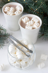 Cocoa with marshmelow in white cups and marshmallows in a glass jar on a white table with spruce branches and garland.