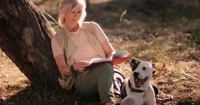Mature woman with dog sitting under tree and reading book