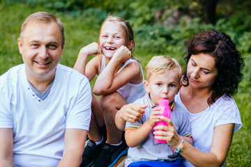 Joyful family with two children. Father, mother and siblings kids having fun outdoors in garden, playing together in summer park. Mom giving son drink from the bottle. Daughter laughing on background.