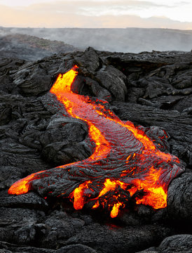 A lava flow emerges from an earth column and flows in a black volcanic landscape, in the sky shows the first daylight - Location: Hawaii, Big Island, volcano "Kilauea"