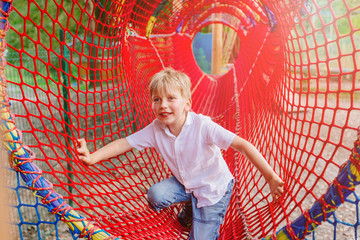 Blond scandinavian school age boy having fun in red net tunnel in playground or amusement park. Holiday friendship and happy childhood concept.