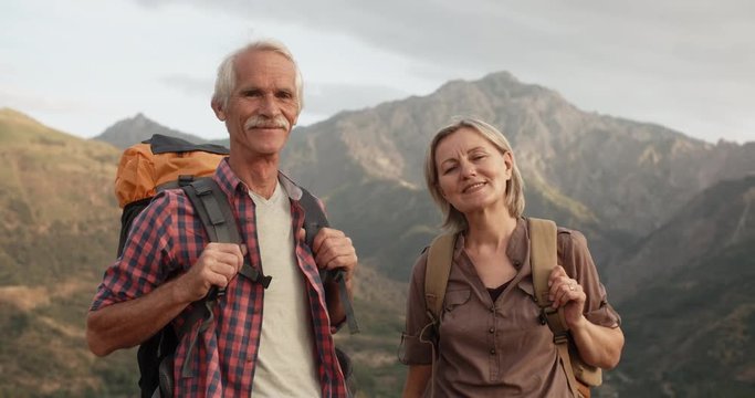 Positive old caucasian couple trekking in mountains with backpacks, enjoying their adventure - portrait shot 4k