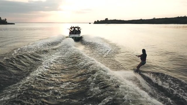 Flying over wakeboarder surfing behind boat. Aerial view of wakeboarding on river at sunset.