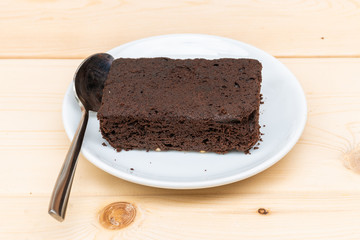 close-up of plain chocolate brownie in white plate with spoon on wooden plank