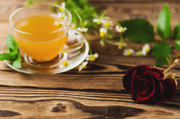 Glass cup with green tea and mint and bunch of chamomile near red rose on sprig on old wooden table. Saint Valentine's Day concept