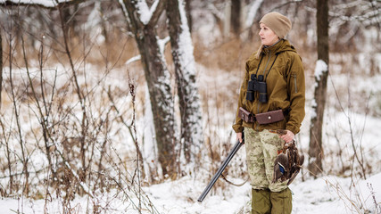 Female hunter in camouflage, armed with a rifle, standing in a snowy winter forest with duck prey