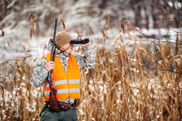 Male hunter in camouflage, armed with a rifle, standing in a snowy winter forest with duck prey © kaninstudio