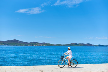 Beautiful female cyclist in white summer closing and hat riding bicycle along stony sidewalk on...