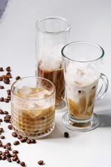 Iced coffee cocktail or frappe with ice cubes and cream served in three different glasses with coffee beans around on white marble table.