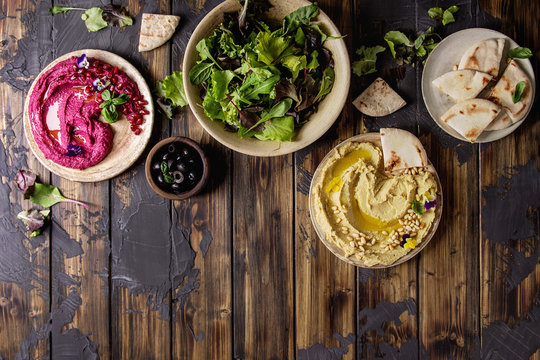 Fototapeta Variety of homemade traditional and beetroot spread hummus with pine nuts, olive oil, pomegranate served on ceramic plates with pita bread and green salad on dark wooden background. Flat lay, space.