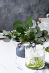 Matcha green tea iced latte or cocktail in glass with ice cubes, matcha powder and jug of milk on white marble table, decorated by green branches. Grey wall at background