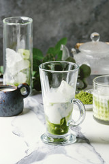 Matcha green tea iced latte or cocktail in three different glasses with ice cubes, matcha powder and jug of milk on white marble table, decorated by green branches. Grey wall at background