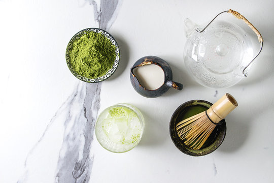 Ingredients for making matcha ice drink. Green tea matcha powder in ceramic bowl, traditional bamboo spoon, whisk, milk, glass teapot, ice cubes over white marble background. Flat lay, space