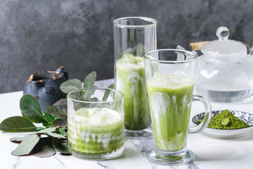Matcha green tea iced latte or cocktail in three different glasses with ice cubes, matcha powder and jug of milk on white marble table, decorated by green branches. Grey wall at background