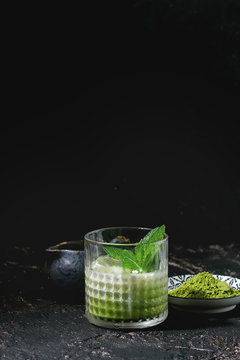 Matcha green tea iced latte or cocktail in glass with ice cubes, mint, matcha powder and jug of milk over dark texture background