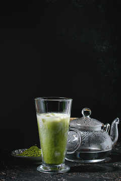 Matcha green tea iced latte or cocktail in tall glass with ice cubes, mint, matcha powder and transparent teapot with hot water over dark texture background