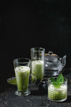 Matcha green tea iced latte or cocktail in three different glasses with ice cubes, matcha powder and transparent teapot over dark texture background