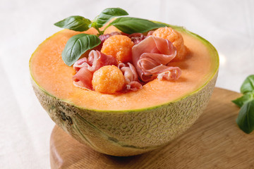 Melon and ham or prosciutto salad served in half of Cantaloupe melon, decorated by fresh basil standing on wooden serving board over white tablecloth. Close up © Natasha Breen
