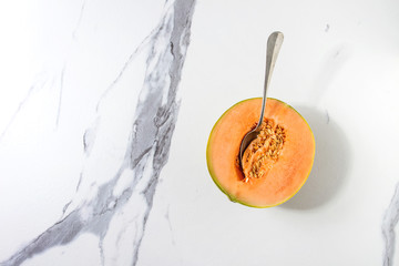 Half of ripe organic Cantaloupe melon with seeds and spoon inside over white marble texture background. Flat lay, space