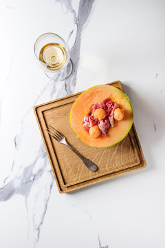 Melon and ham or prosciutto salad served in half of Cantaloupe melon on wooden square serving board over white marble background with glass of white wine and fork. Flat lay, space