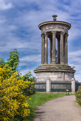 Edinburgh, Scotland, UK - June 13, 2012: Cylindrical gray stone memorial for Donald Stewart on Calton Hill under blue cloudy sky. Some yellow flowers and green vegetation.