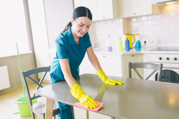 Positive female cleaner looks down and leans to table. She cleans it. Girl works in studio apartment in kitchen. She wears blue uniform and yellow gloves.