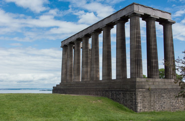Fototapeta na wymiar Edinburgh, Scotland, UK - June 13, 2012: Free standing National Monument is row of pillars under blue sky with white clouds. North Sea inlet in back, green grass in front.