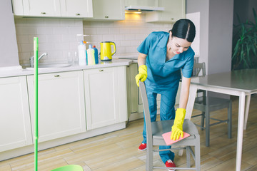 Concent5rated woman bows down to chair and clean it with rag. She works in kitchen. Girl wears blue uniform and yellow gloves.