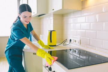 Cheerful cleaner works at stove. She cleans it with orange rag. Girl wears yellow gloves and blue uniform. She looks happy.