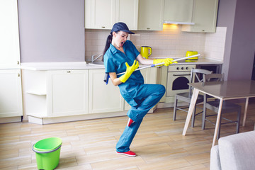 Funny worker dancing in kitchen. She pretends playing on guitar. But girl has mop in hands in stand of that. She is rocking and relaxing.