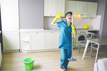 Fantastic woman stands in kitchen and holds mop in hands. She waves with hands and singing out loud. Cleaner has rest.