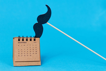 Fake mustache on the stick and paper calendar with November 2018 open page on blue background - symbol of Movember, event to raise awareness prostate and testicular cancer and men's suicide