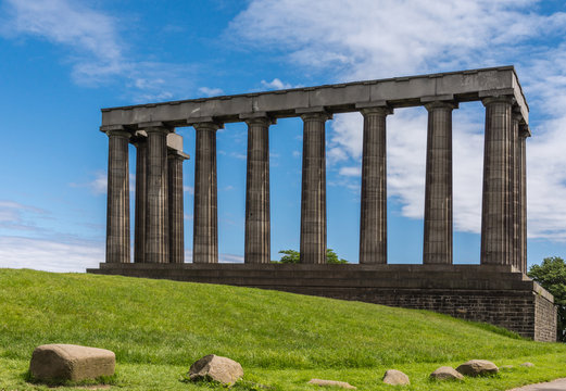 Edinburgh, Scotland, UK - June 13, 2012: National Monument is series of dark brown pilars against blue sky with white clouds. Green grass up front.