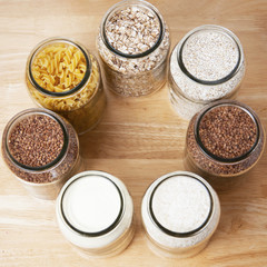 Various uncooked cereals, grains, and pasta for healthy cooking in glass jars on wooden table. Top view. Clean eating, balanced dieting food. Square image.
