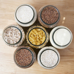 Various uncooked cereals, grains, and pasta for healthy cooking in glass jars on wooden table. Top view. Clean eating, balanced dieting food. Square image.