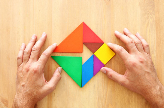 man's hand holding a missing piece in a square tangram puzzle, over wooden table