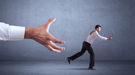 Young miniature businessman running from a big hand with blueish grey background