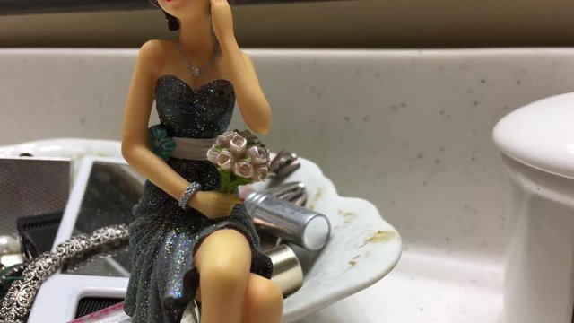 Plastic pretty woman in dress sitting in bathroom with jewelry and other various items in background. 