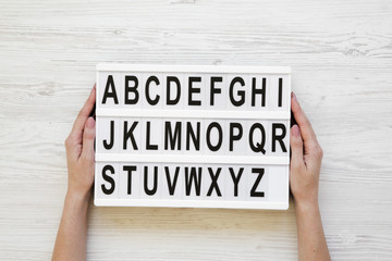 Female hands hold modern board with english alphabet on lightbox over white wooden background, from above. Closeup.
