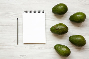Whole avocados, blank notepad and pencil on white wooden table, top view. Flat lay, overhead, from above. Space for text.