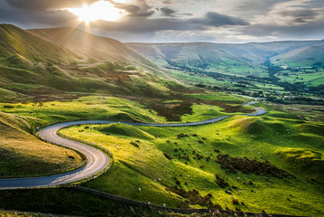 Sunset at Mam Tor, Peak District National Park, with a view along the winding road among the green hills down to Hope Valley, in Derbyshire, England. - obrazy, fototapety, plakaty
